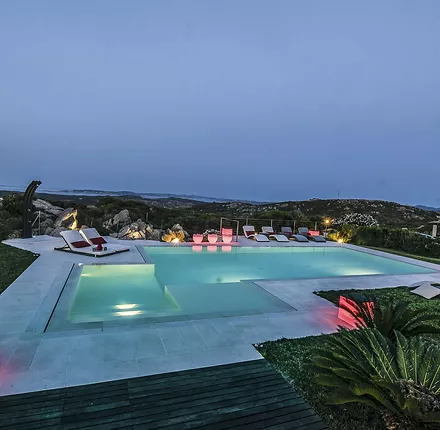 Piscina privata a sfioro Trilogy con scala e area relax interna, realizzata in Sardegna - piscina illuminata Piscina privata a sfioro Trilogy con scala e area relax interna, realizzata in Sardegna - piscina illuminata